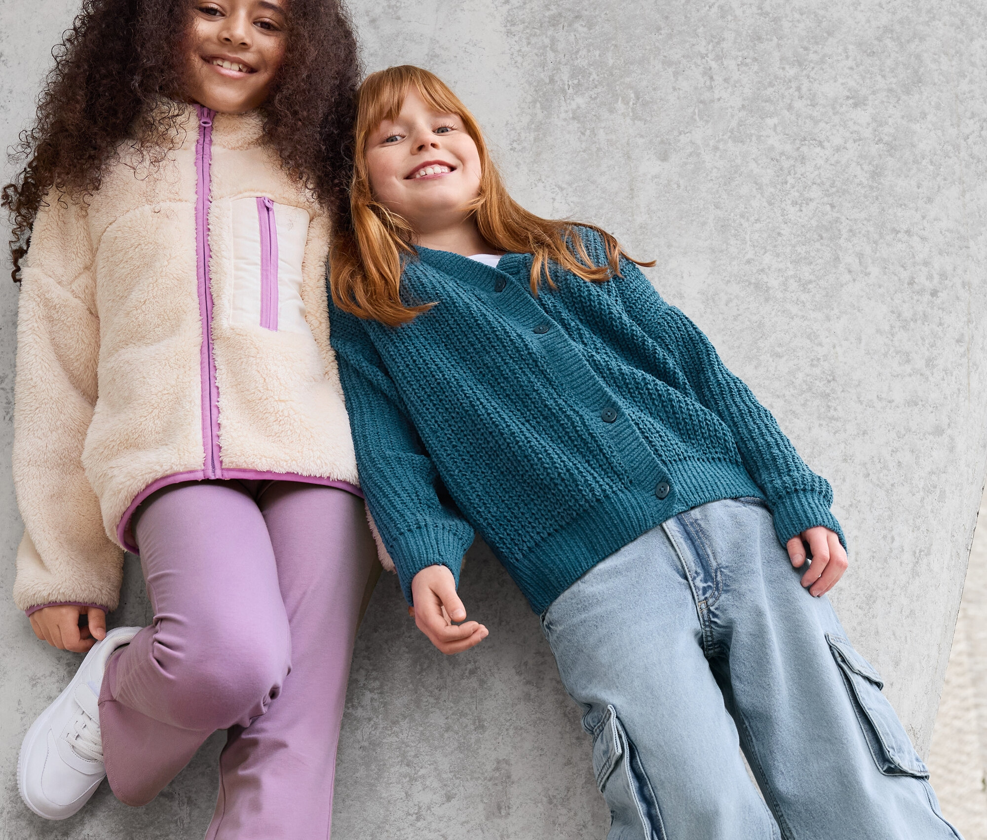 Dvě dívky se opírají o betonovou zeď. Dívka vlevo má na sobě bundu z umělé kožešiny, fialové kalhoty a bílé tenisky. Dívka vpravo má na sobě zelený kardigan a modré kalhoty.