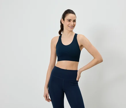 Žena v tmavě modré sportovní podprsence a legínách pózuje na bílém pozadí.