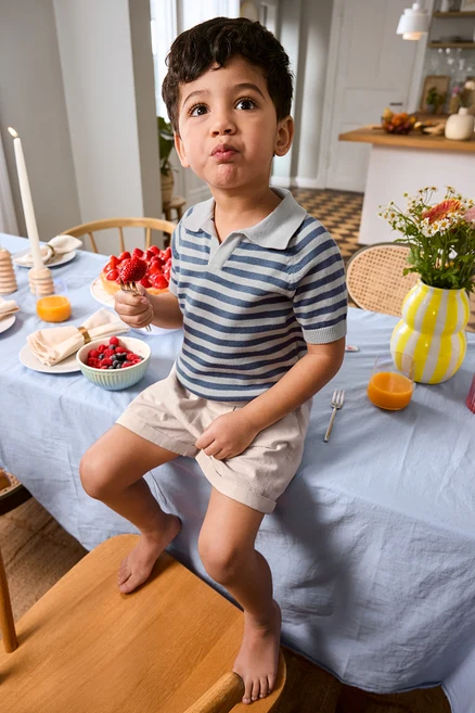 Chlapec sedí na okraji stolu s jahodou na vidličce, má na sobě Kinder-Shorts a Kinder-Strick-Poloshirt. Na stole váza a dortový podnos.