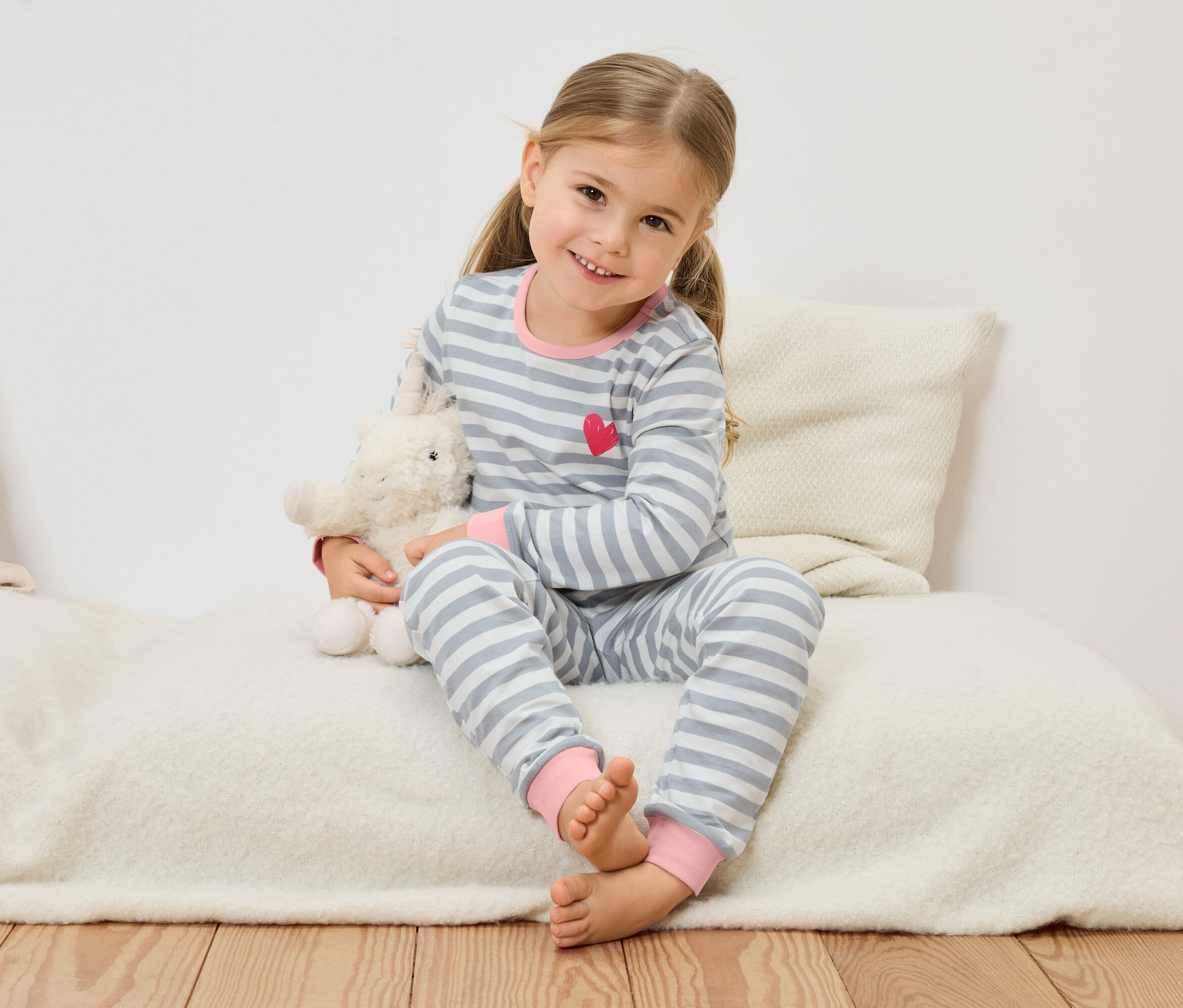 Malá dívka s blond vlasy sedí na polštáři a drží plyšovou ovečku.