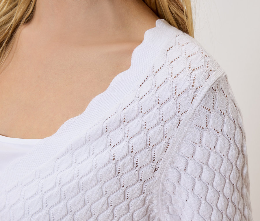 Detailní záběr na ženu s blond vlasy v bílém pleteném kardiganu s děrovaným vzorem na bílém topu.