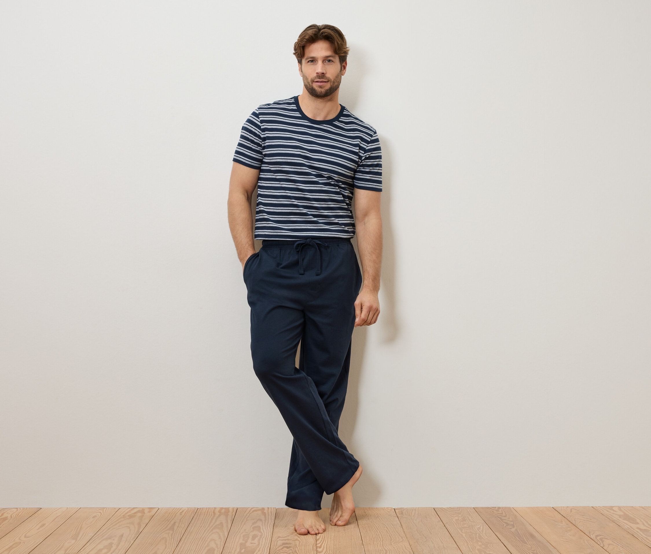 Muž se opírá o zeď, má na sobě modrobílé pruhované tričko a tmavě modré pyžamové kalhoty.