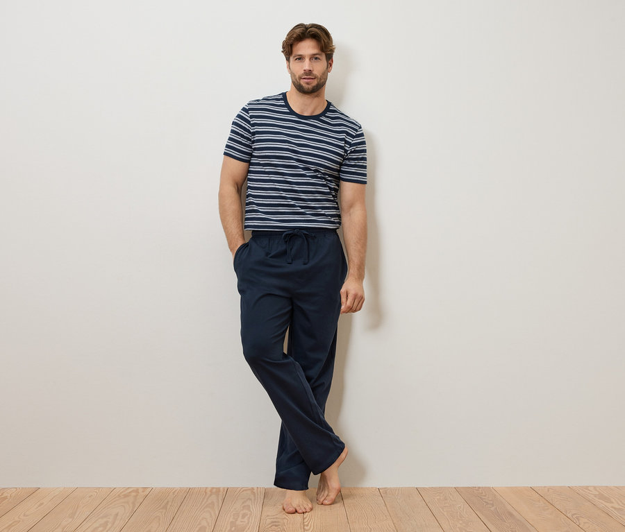 Muž se opírá o zeď, má na sobě modrobílé pruhované tričko a tmavě modré pyžamové kalhoty.
