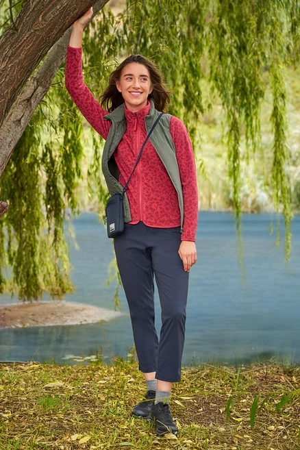 Žena se opírá o strom u jezera, má na sobě červenou fleecovou bundu, tmavě modré kalhoty a khaki vestu »packable« Hybrid-Weste.