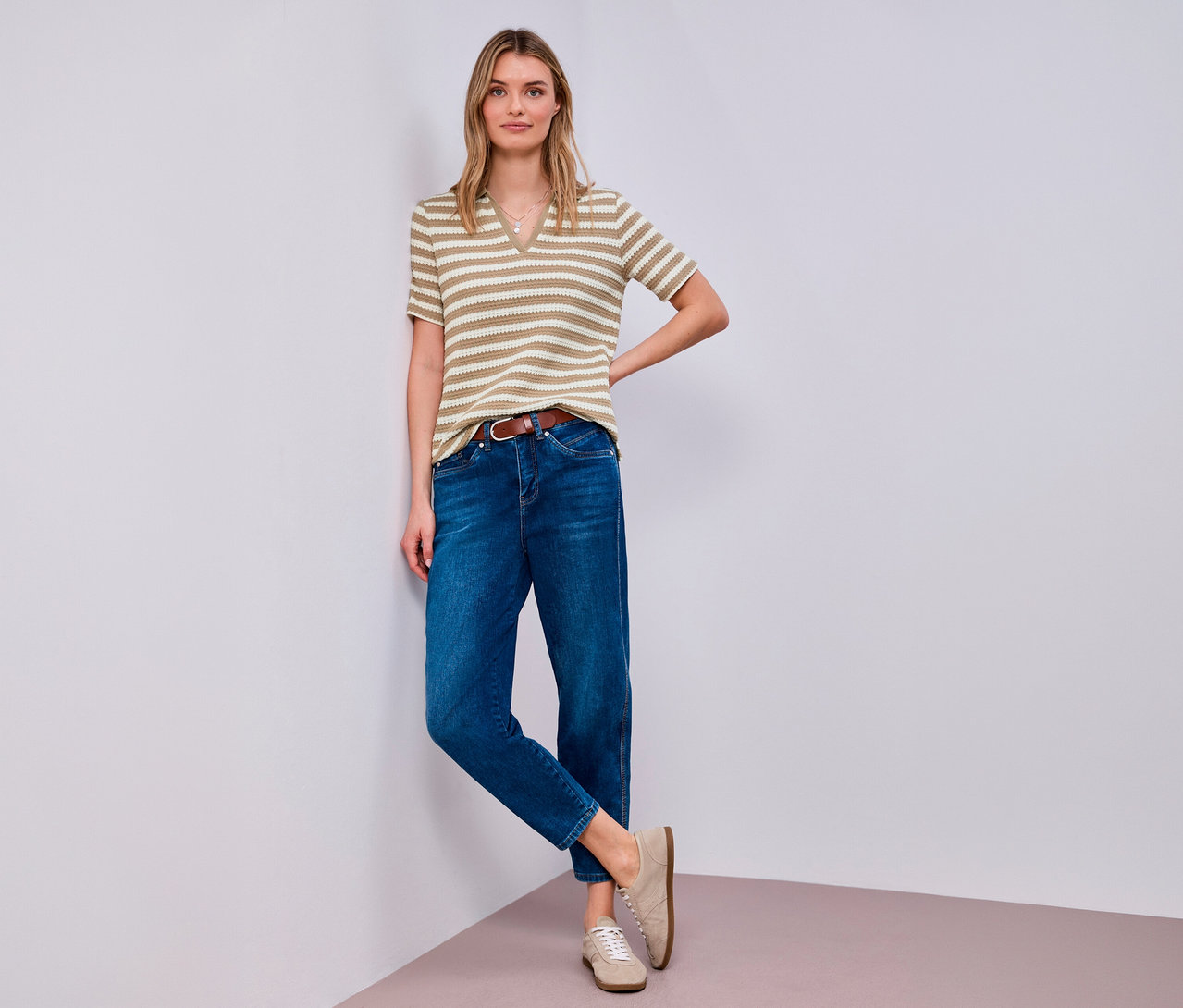 Žena pózuje v úpletovém tričku Street One, džínách, hnědém pásku a béžových botách v rohu.