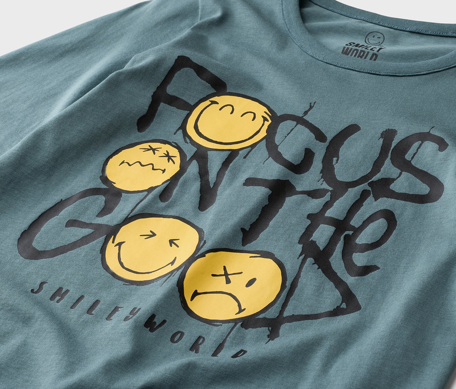 Detailní záběr na petrolejové tričko s dlouhým rukávem Smiley World s nápisem „Focus on the Good“ a žlutými smajlíky.