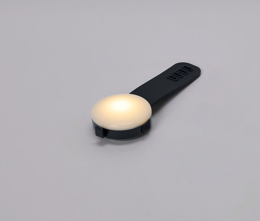 Detailní záběr malé, kulaté, svítící lampy s černou rukojetí na šedém pozadí.