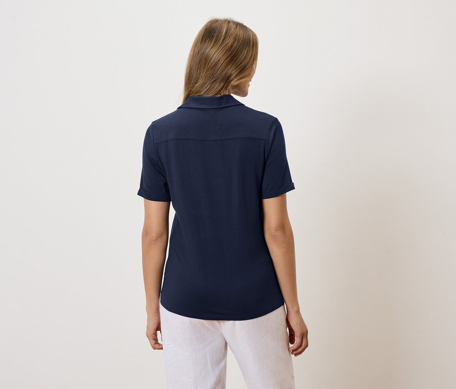 Pohled zezadu na blond ženu v tmavě modré polokošili a bílých kalhotách.