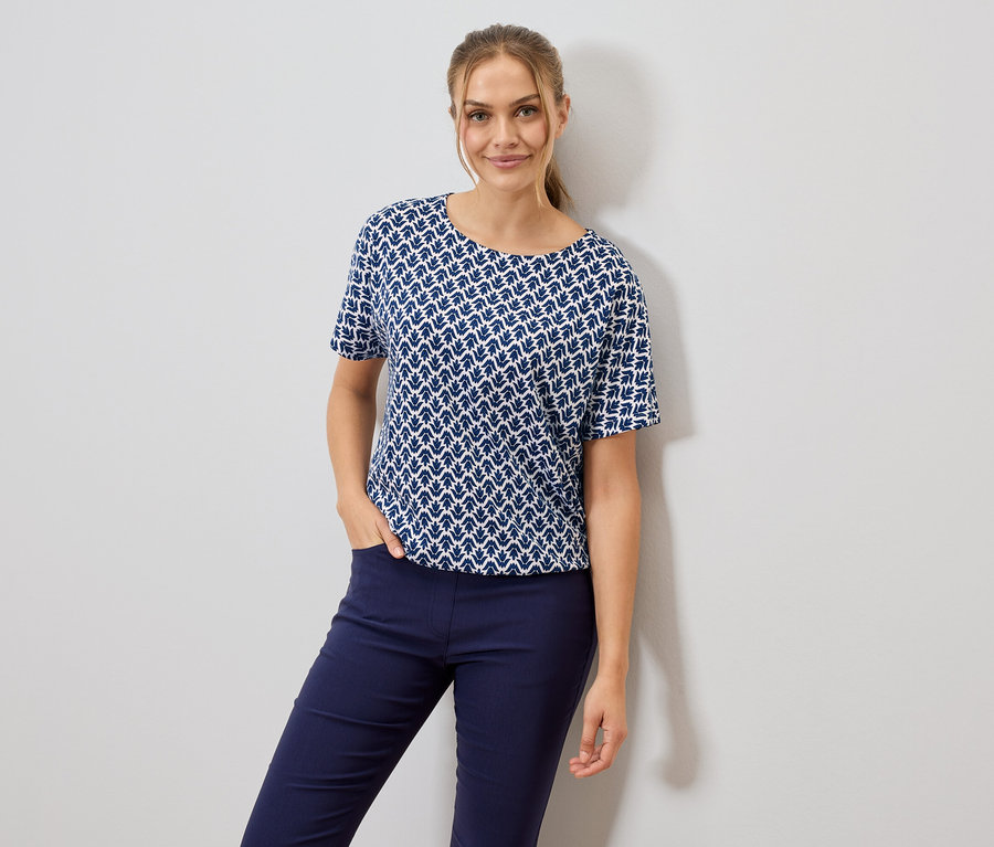 Žena v modro-bílém vzorovaném tričku a modrých kalhotách, s jednou rukou v kapse.