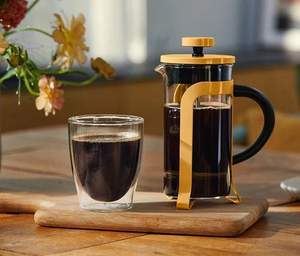 Dvoustěnná sklenice černé kávy a žlutý French Press plný kávy stojí na dřevěné desce. V pozadí je váza s květinami a talířek s sušenkou.