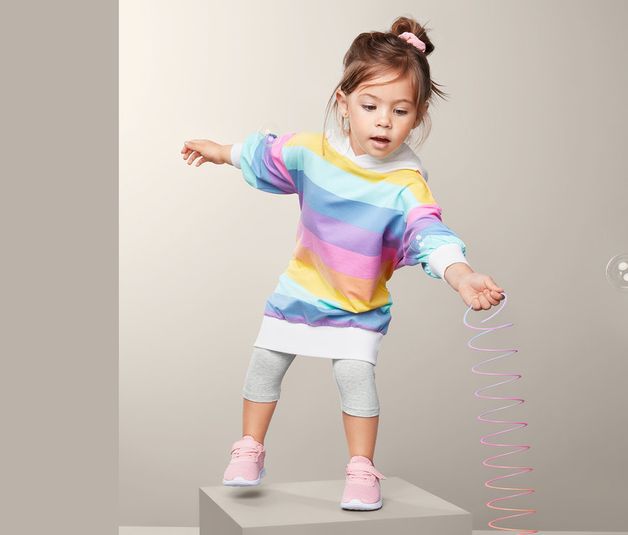 Dívka stojí na šedé kostce a drží natahovací hračku. Má na sobě duhové šaty a šedé legíny, 2 ks.