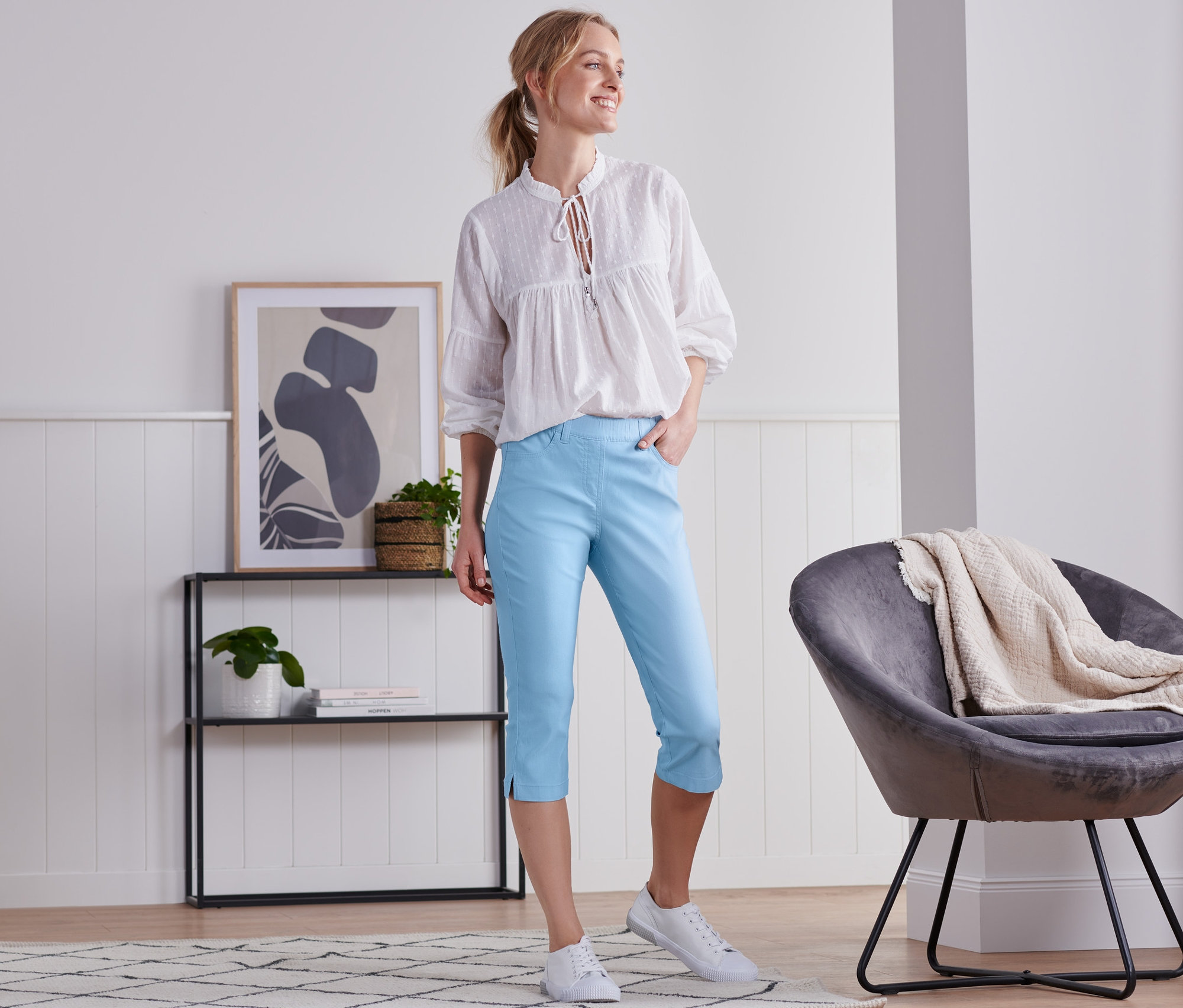 Žena stojí v místnosti a má na sobě světle modré tříčtvrteční kalhoty a bílou blůzu.