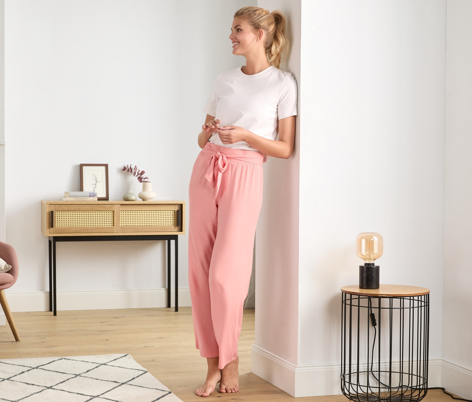 Blondýna se usmívá a opírá se o zeď v růžových pohodlných kalhotách.