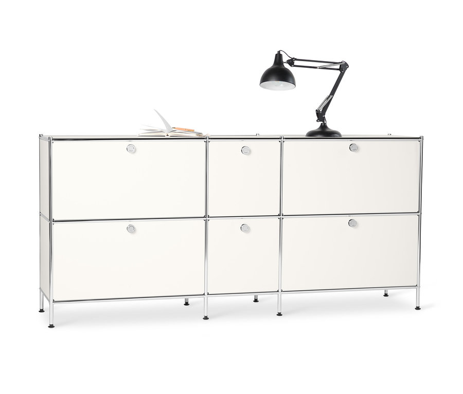 Bílá kovová odkládací skříňka CN3 se 6 výklopnými dvířky. Na horní straně leží otevřený zápisník a stojí černá lampa.