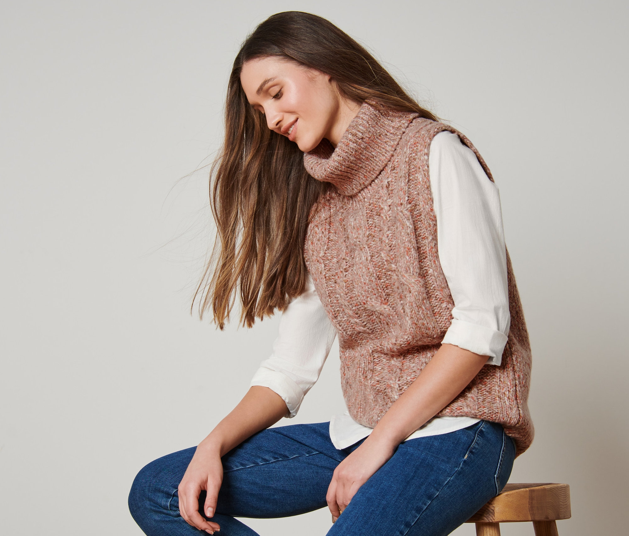 Žena sedí na stoličce a má na sobě pletenou vestu s copánkovým vzorem.