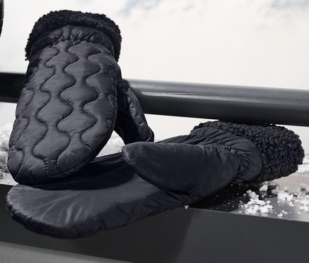 Černé prošívané palčáky a lyže opřené o zábradlí s horami v pozadí.