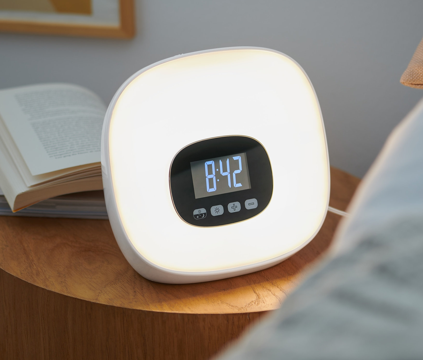 Na nočním stolku stojí světelný budík s rádiem vedle knih. Na displeji budíku svítí čas 8:42.