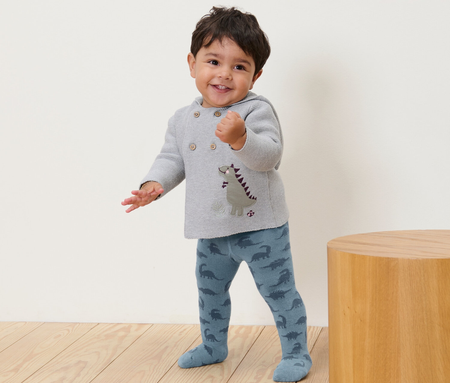 Usmívající se batole stojí a má na sobě modré punčocháče se vzorem dinosaurů.