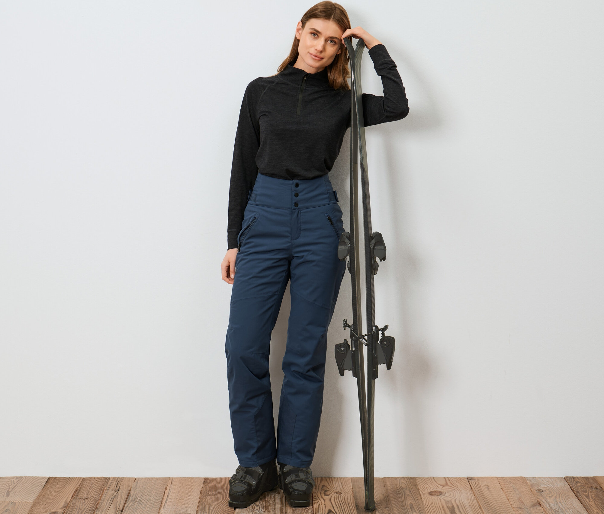 Žena opírající se o lyže má na sobě modré lyžařské kalhoty s vysokým pasem.