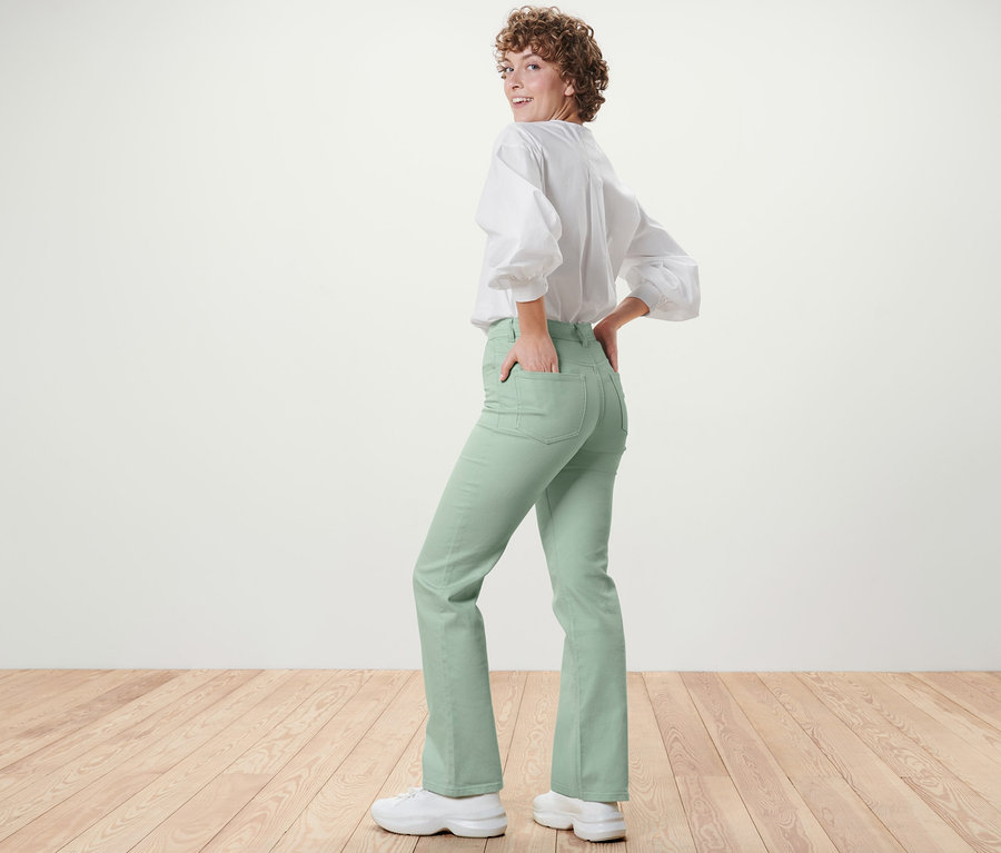 Žena se dívá přes rameno a pózuje v mátově zelených džínách bootcut střihu Lou a bílé halence.