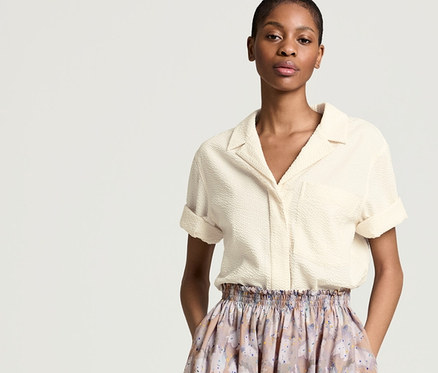 Žena stojí a má ruce v kapsách sukně. Má na sobě blůzu NAH/STUDIO Seersucker z nebělené bavlny a sukni NAH/STUDIO z biobavlny.