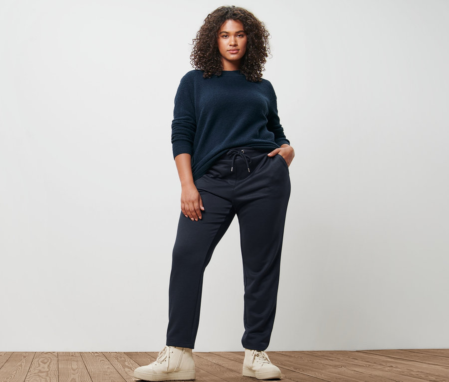 Žena pózuje v pleteném svetru a tmavě modrých joggingových kalhotách.
