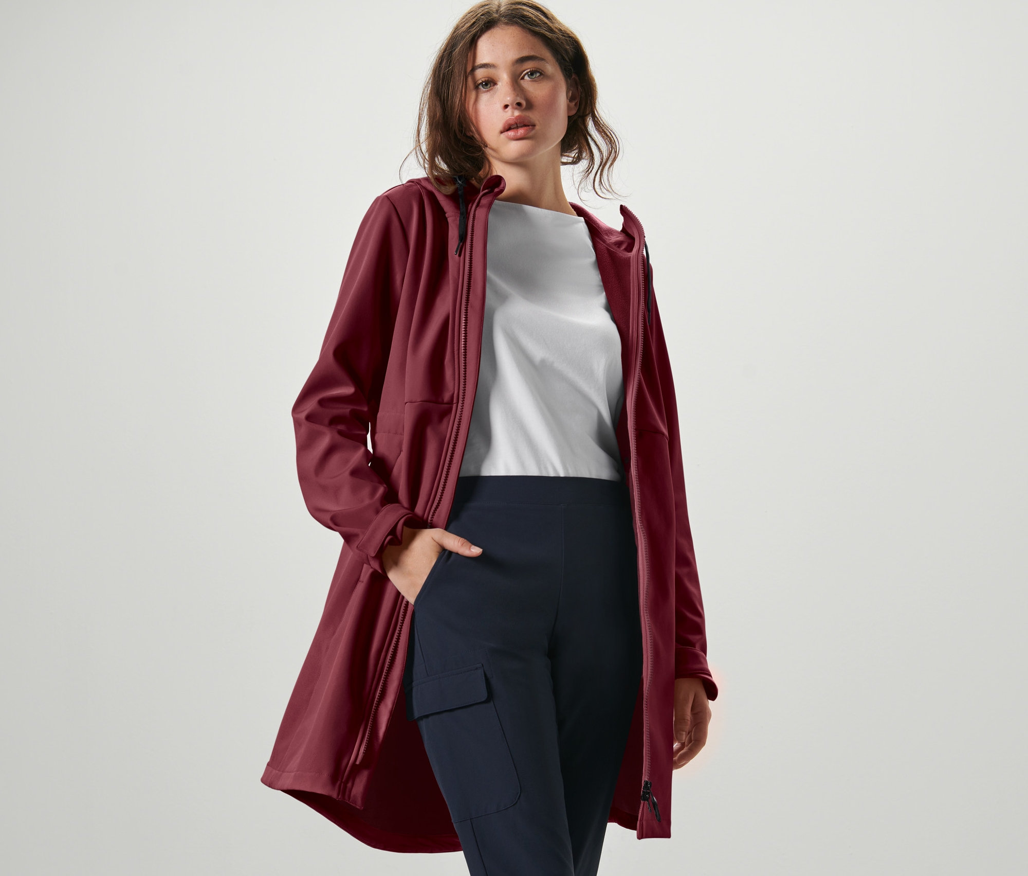 Žena pózuje v softshellovém kabátu vínově červené barvy, bílém tričku a tmavě modrých kalhotách.