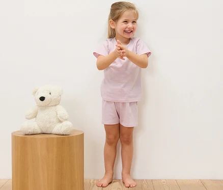 Usmívající se dívka tleská v krátkém dětském pyžamu (2 ks) s proužky. Vlevo sedí plyšový medvěd na dřevěném stolku.