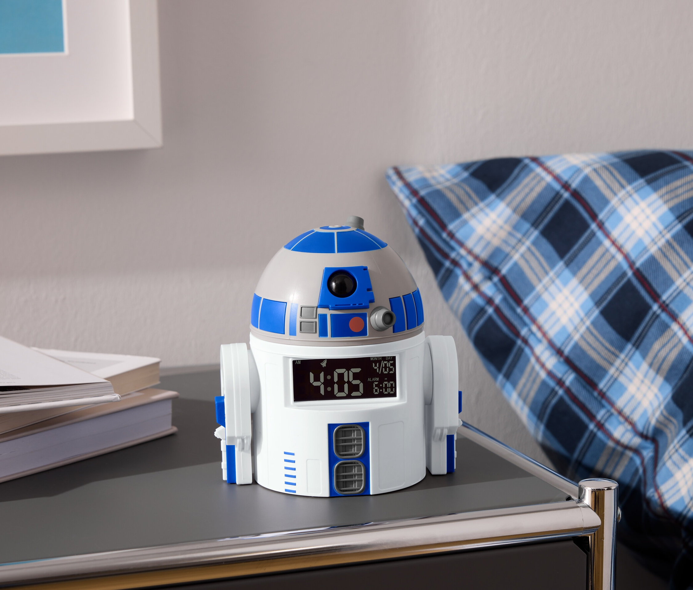 R2-D2 budík stojí na nočním stolku vedle knih a modro-bílé kostkované podušky.