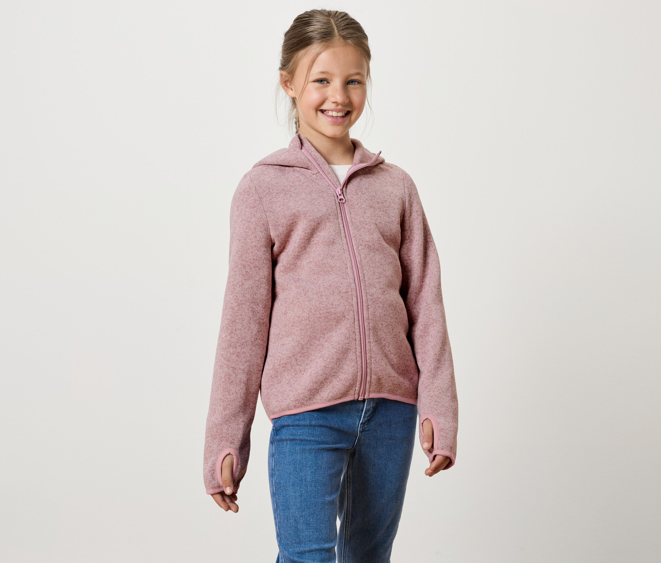 Usmívající se dívka má na sobě růžovou mikinu na zip s kapucí a modré džíny.