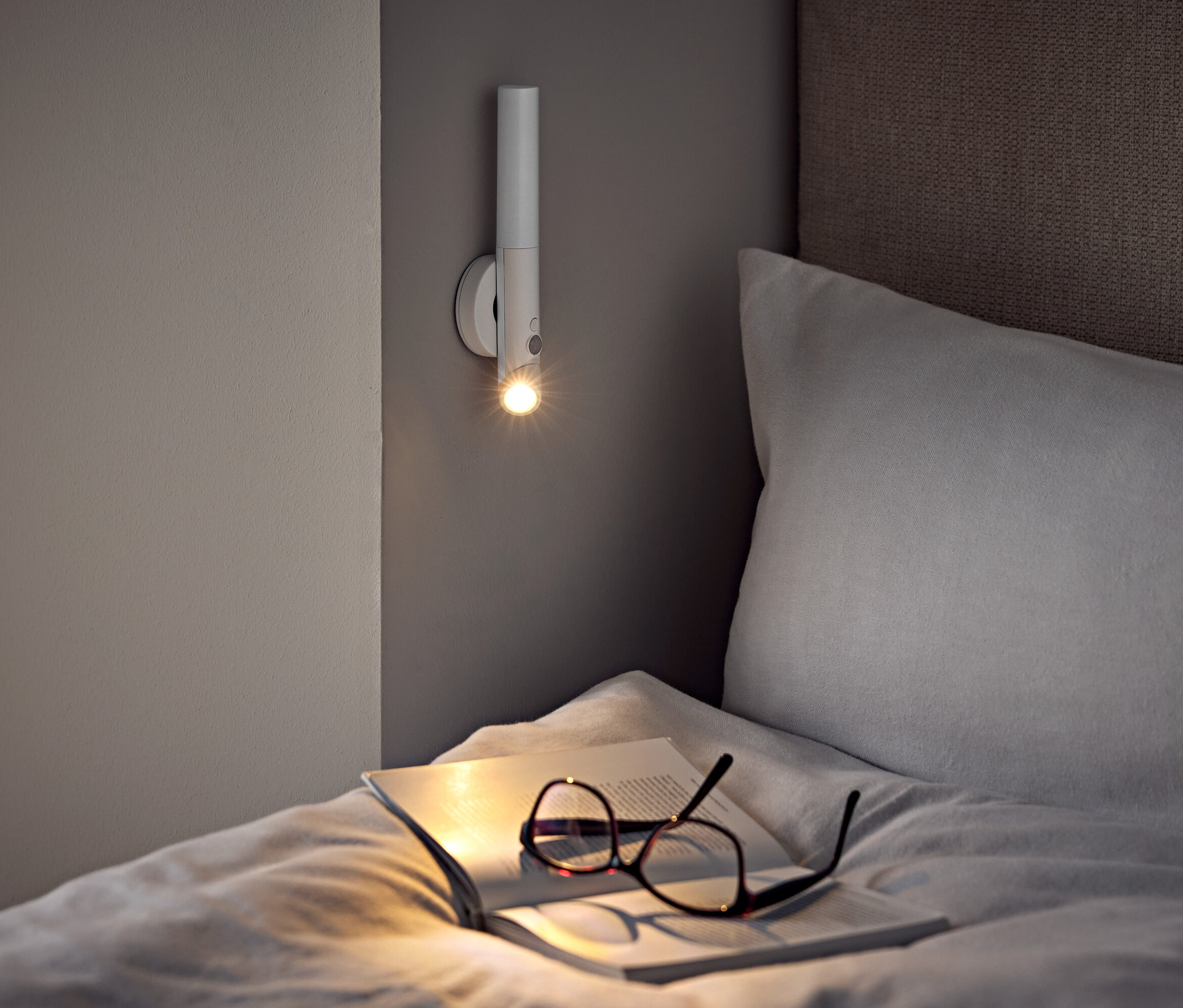 Na posteli leží brýle a otevřená kniha pod nástěnnou lampou.