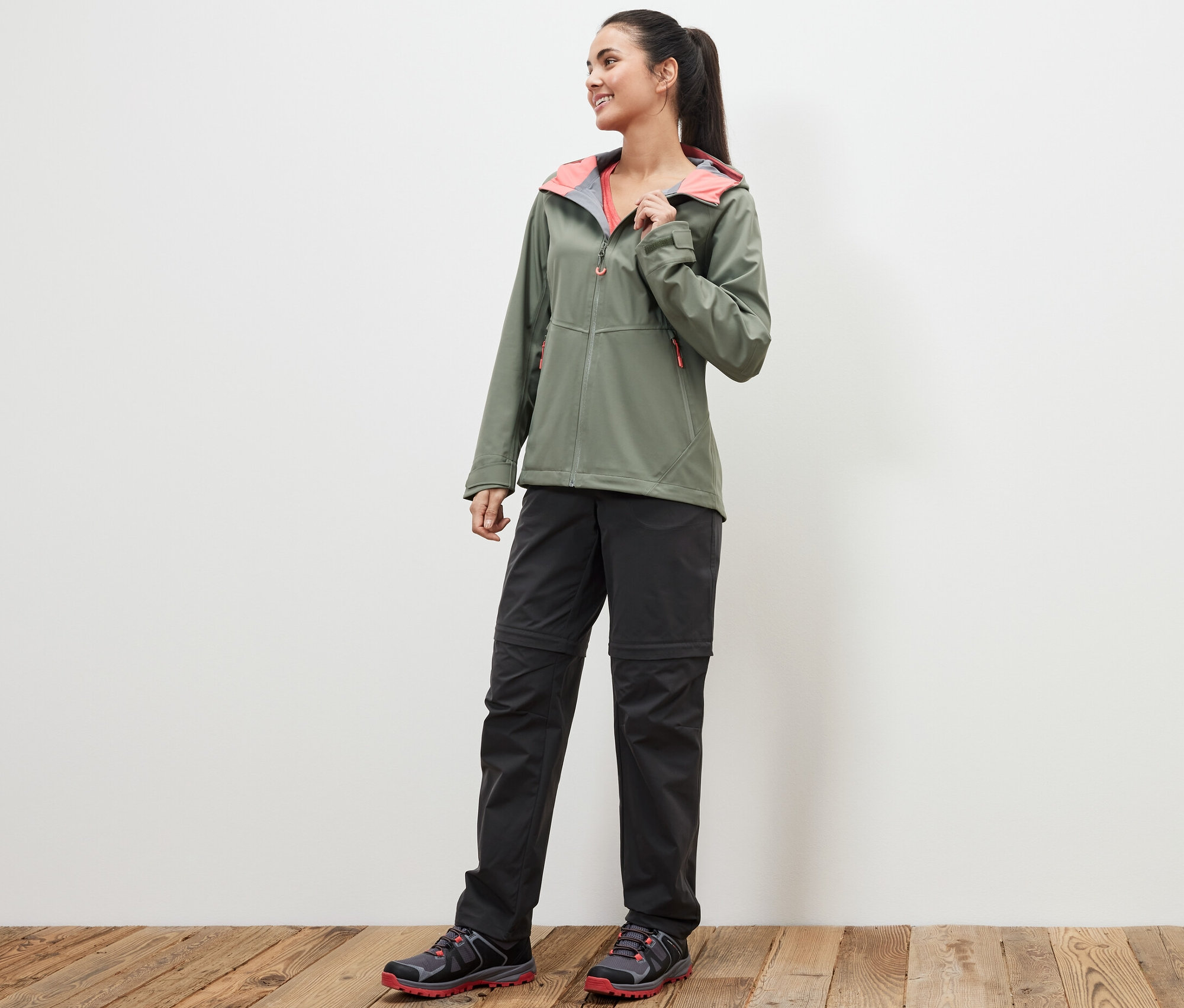 Žena stojící na dřevěné podlaze má na sobě zelenou bundu a černé funkční kalhoty 2 v 1.