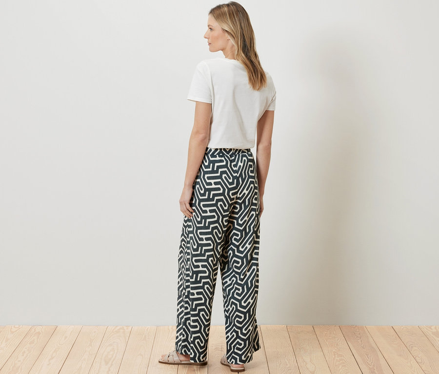 Žena stojí zády k fotoaparátu a má na sobě bílé tričko a navlékací kalhoty v mačkaném vzhledu s potiskem.