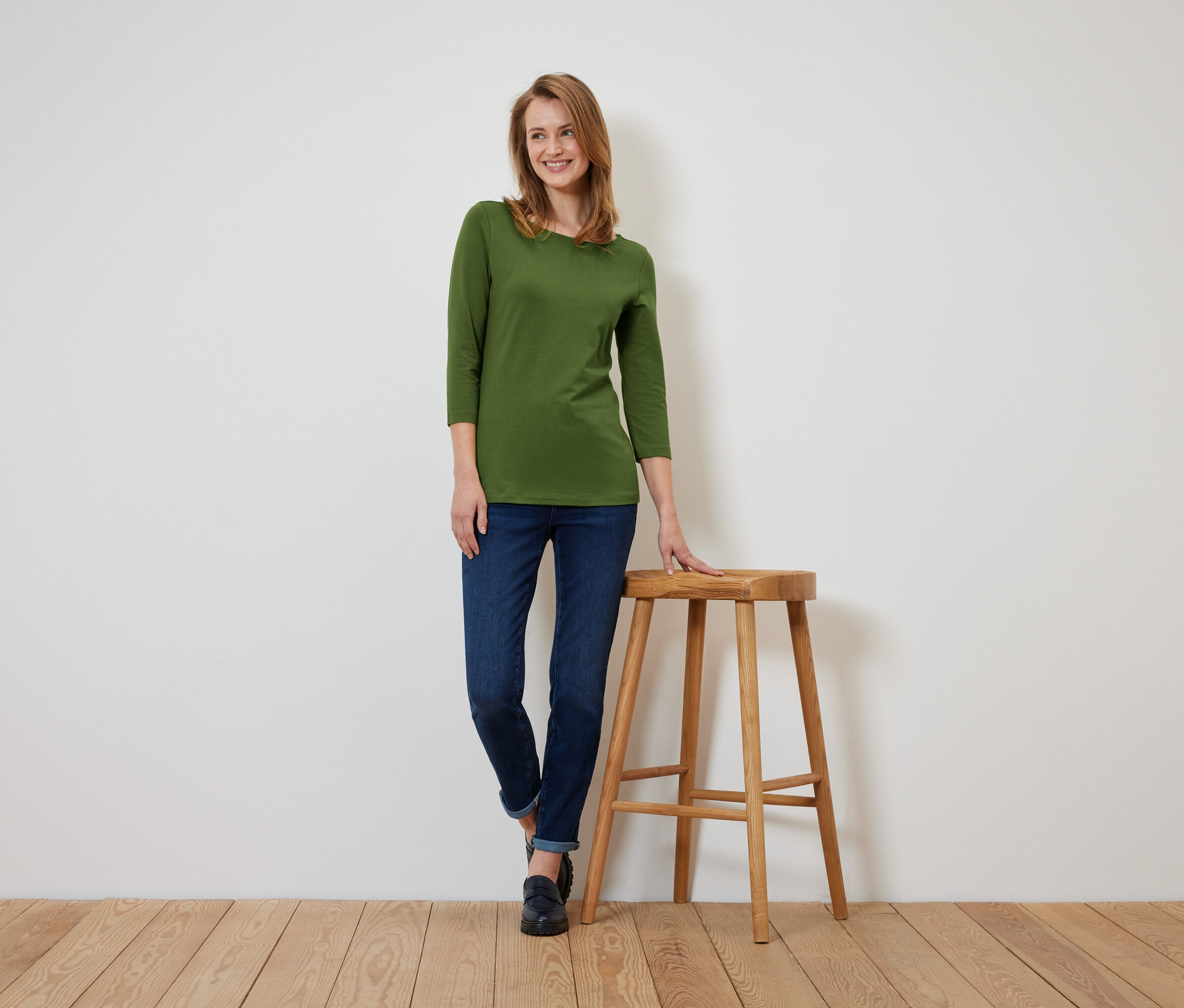 Žena v olivově zeleném tričku se třemi čtvrtinami rukávů a tmavě modrých džínách stojí a opírá se o dřevěnou stoličku.