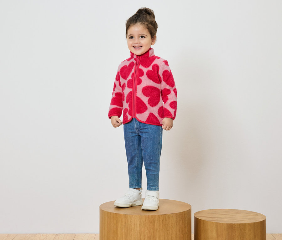 Usmívající se holčička stojí na dřevěném podstavci a má na sobě růžovou fleecovou bundu se vzorem červených srdcí, džíny a bílé tenisky.
