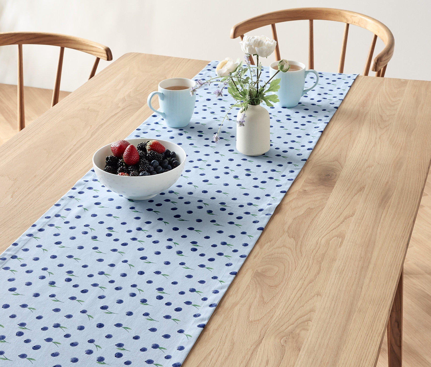 Na stole leží běhoun na stůl s borůvkovým vzorem, na němž stojí mísa s ovocem, dvě modré šálky a váza s květinami.