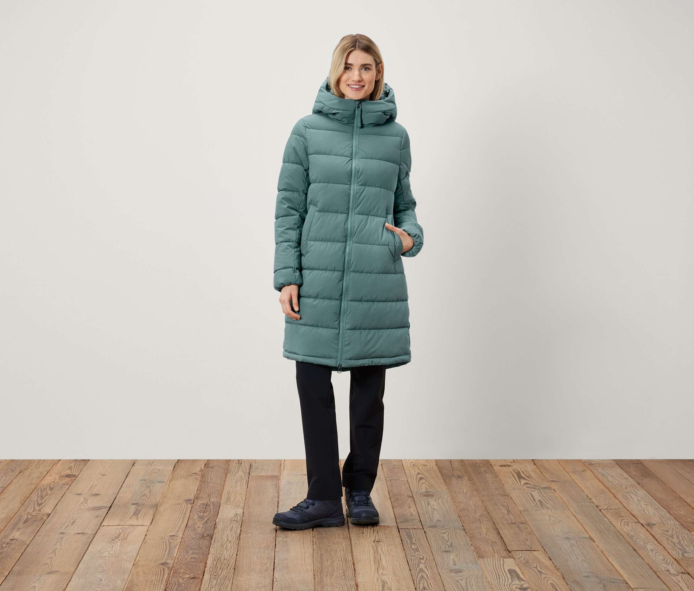 Žena stojí a má na sobě dlouhý prošívaný kabát s kapucí, černé kalhoty a boty.