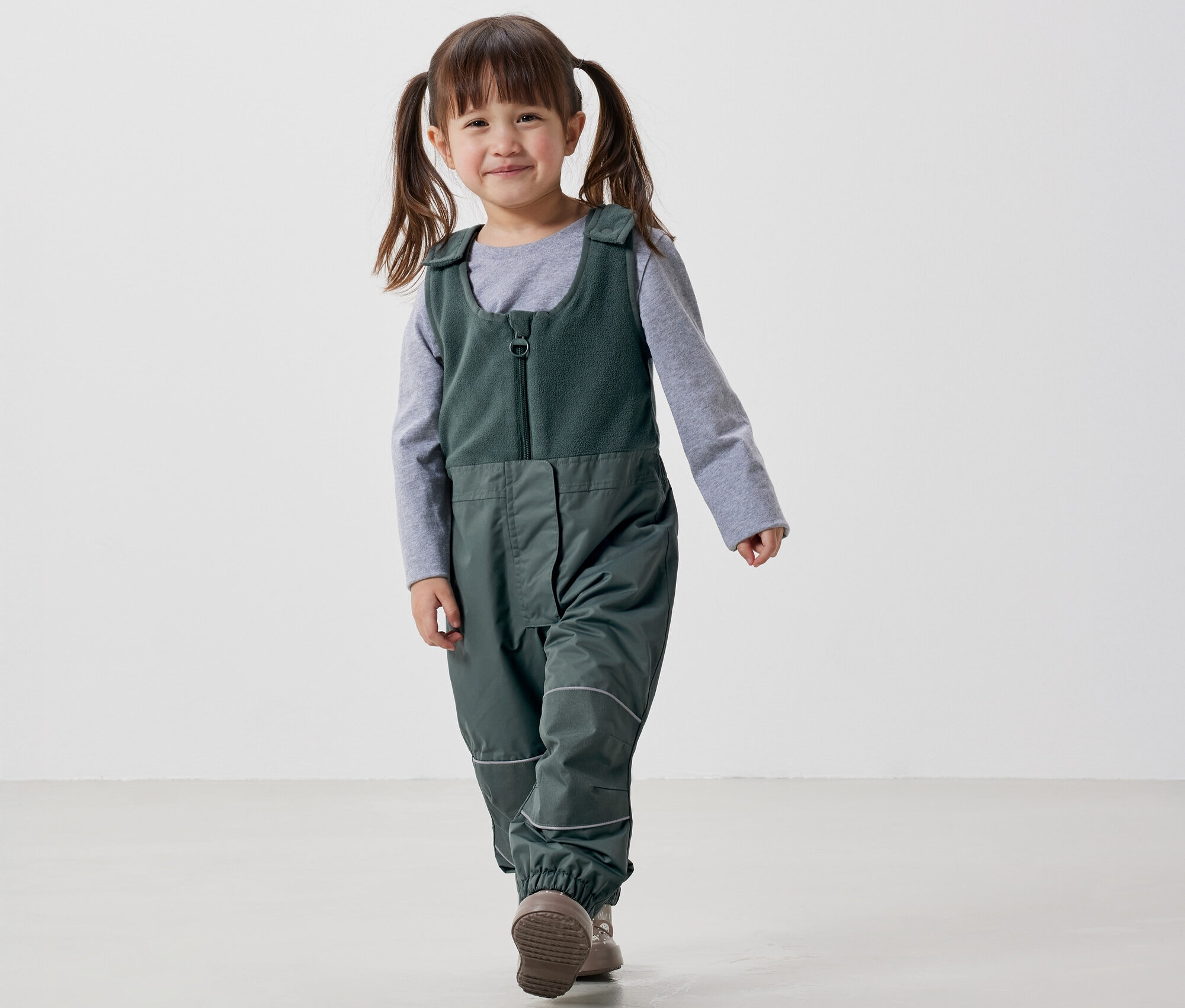 Usmívající se holčička s culíky kráčí v zelených lacláčích a šedém tričku po bílém pozadí.