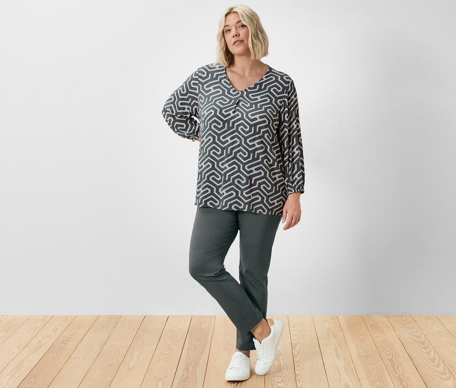 Žena pózuje ve studiu v tmavě zelených strečových kalhotách a halence v mačkaném vzhledu.