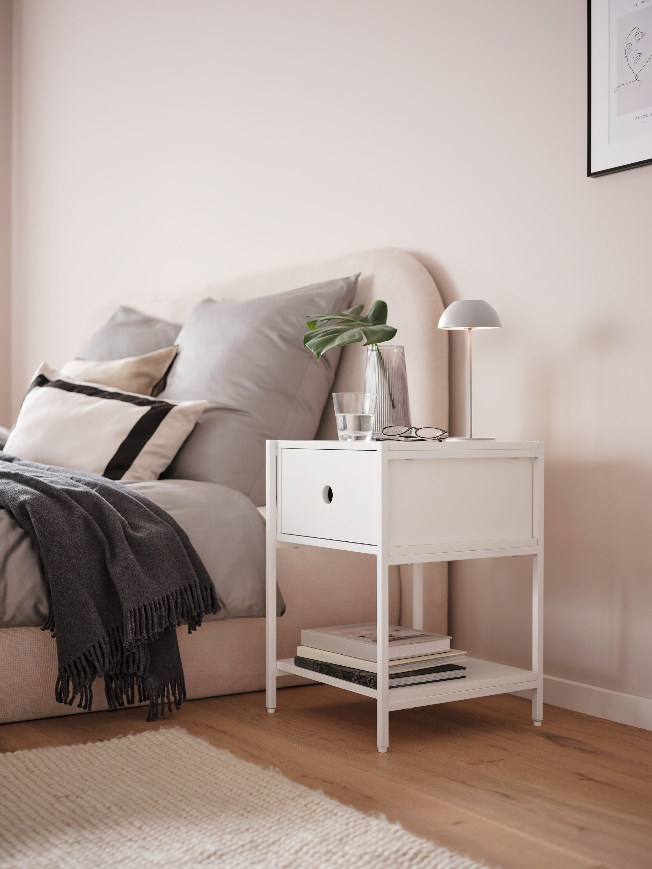 Šedá bateriová stolní lampa s LED na bílém nočním stolku vedle postele.