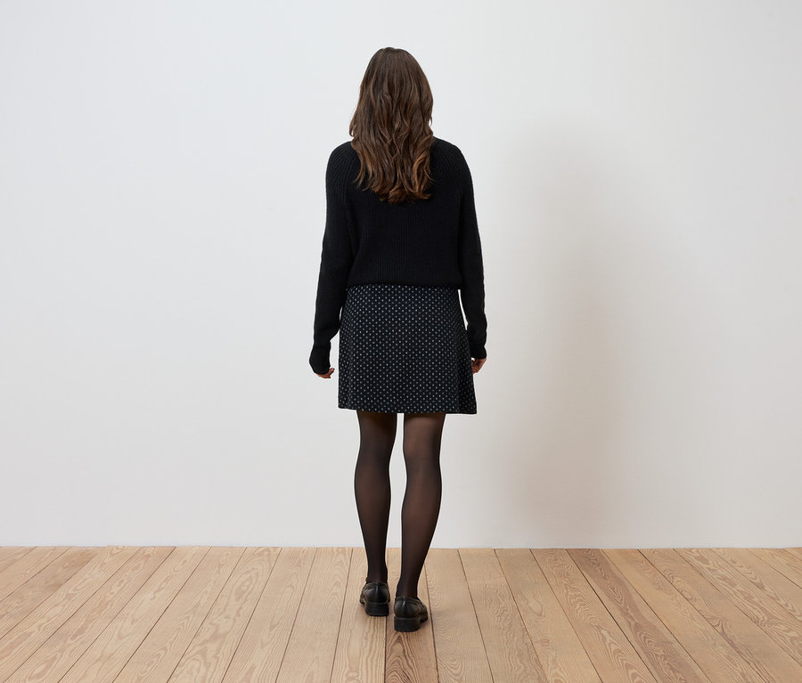 Žena stojí zády k fotoaparátu a má na sobě černý svetr, vzorovanou sukni a černé punčochy.