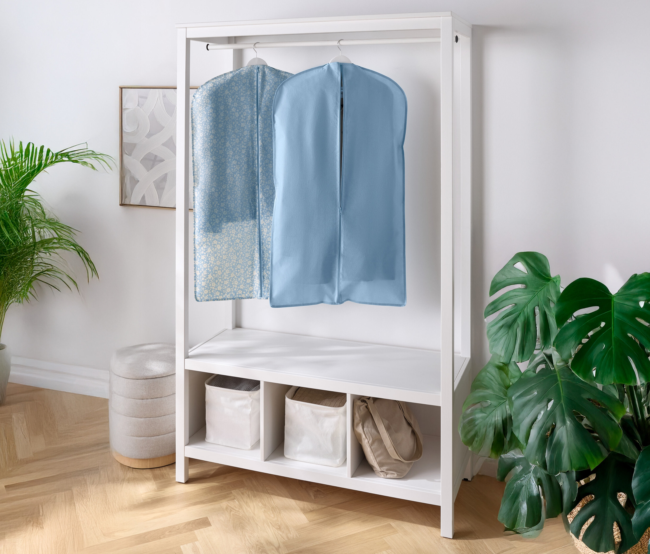 Dva obaly na oblečení visí na bílém šatníku s policemi a přihrádkami pod ním. Vlevo a vpravo je pokojová rostlina.