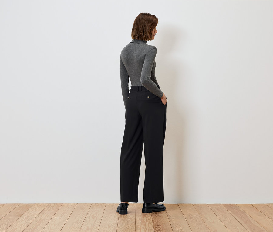 Zezadu je vidět žena s rukama v kapsách černých kalhot a šedým rolákem.