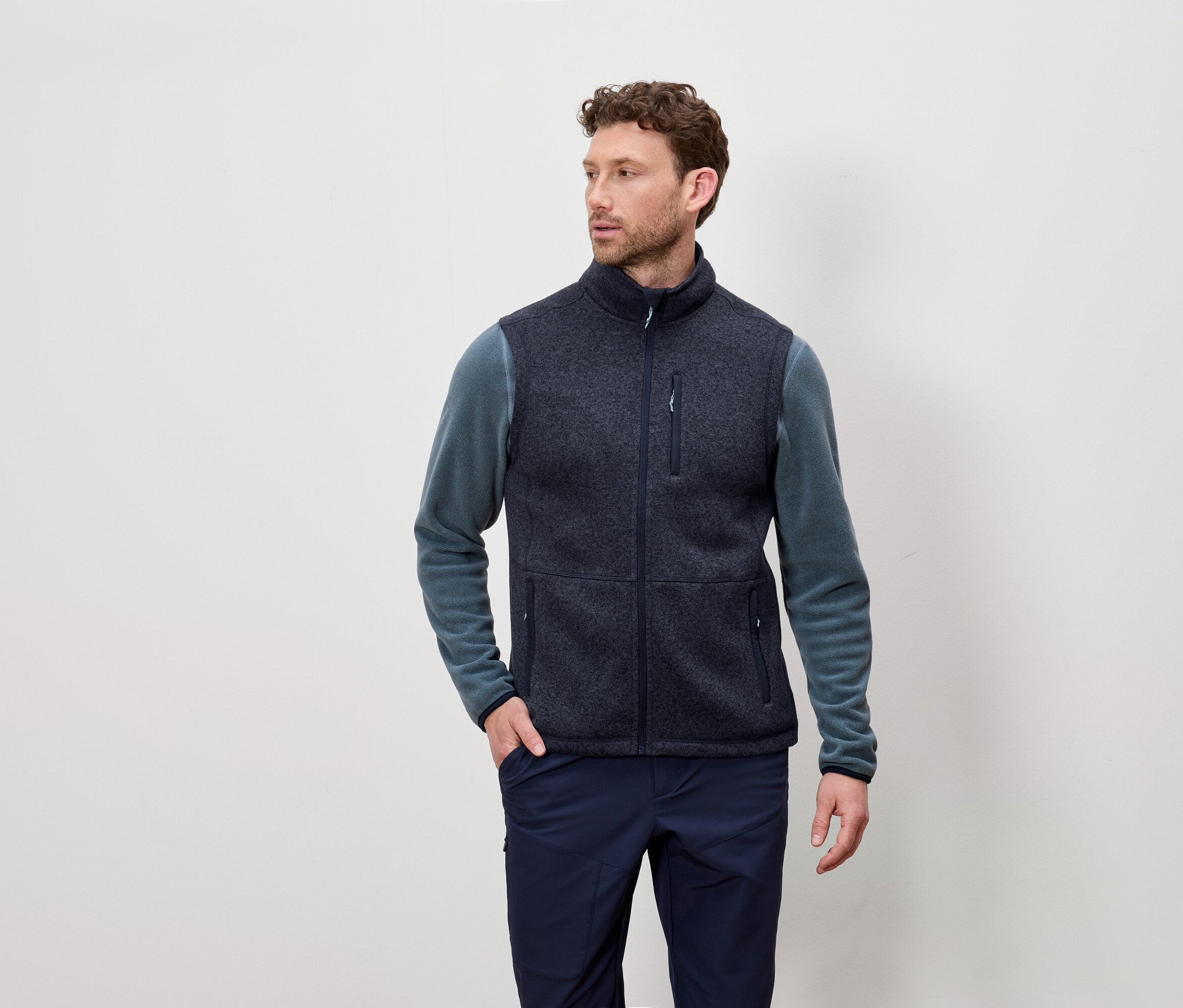 Muž s krátkými kudrnatými vlasy má na sobě tmavě modrou fleecovou vestu a modrozelené tričko s dlouhým rukávem a pózuje na světlém pozadí.