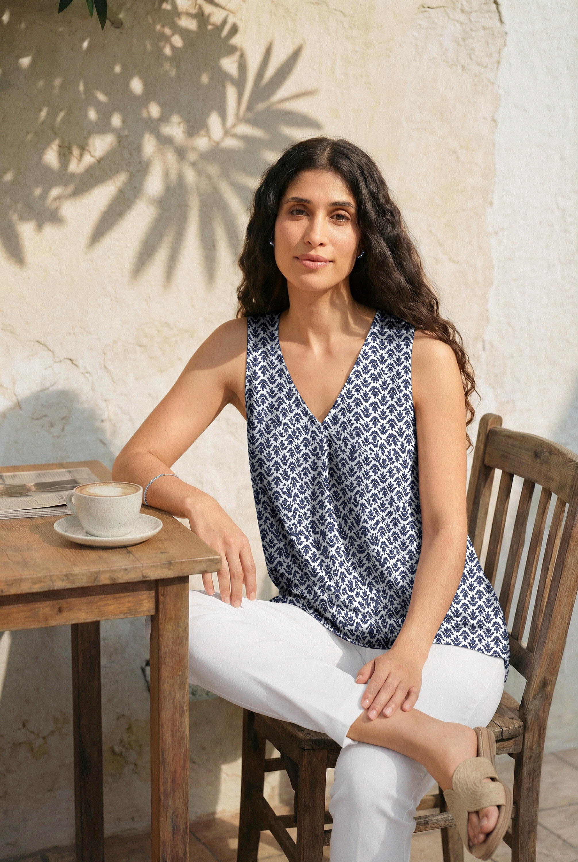 Žena sedí u stolu s náušnicemi a tenisovým náramkem. Má na sobě top s potiskem, bílé 7/8-Stretchhose a pantofle.