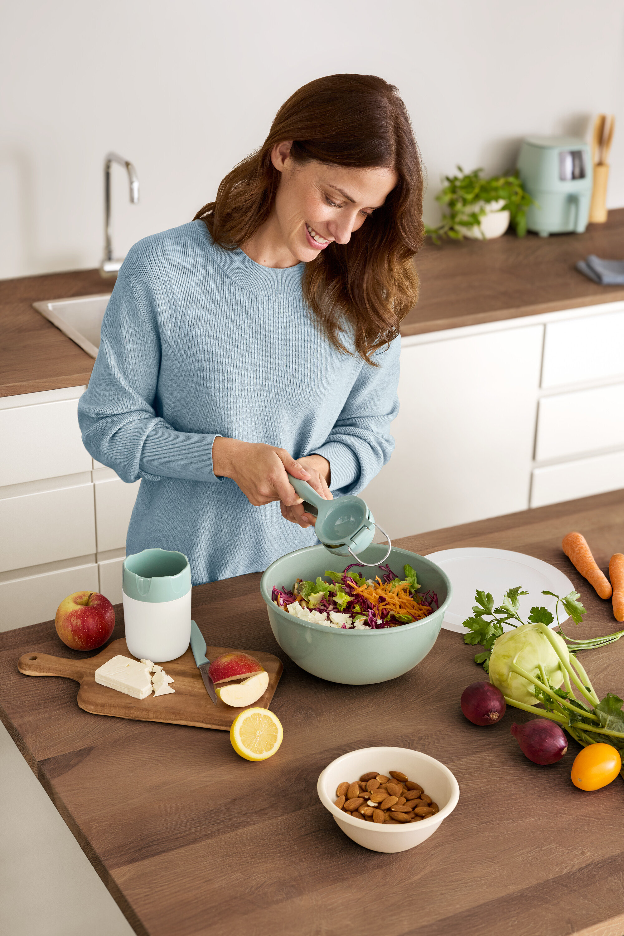 Žena lisuje citron do mísy na salát. Na stole leží jablko, sýr, mandle a zelenina.