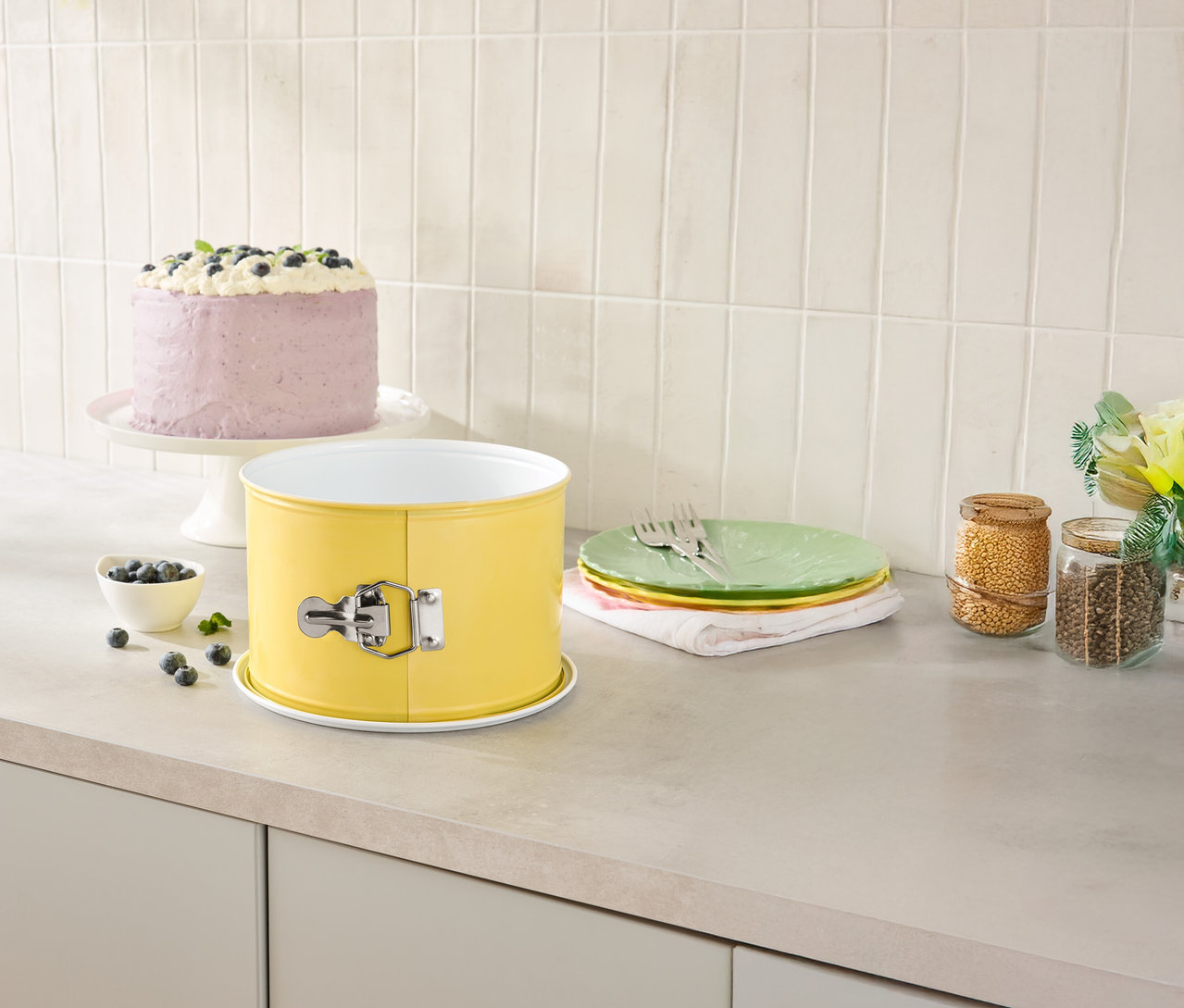 Na kuchyňské lince stojí žlutá forma na pečení, dort na stojanu, talíře a sklenice na potraviny.