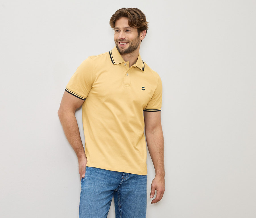 Muž ve žluté polokošili a džínách stojí před světlou zdí.