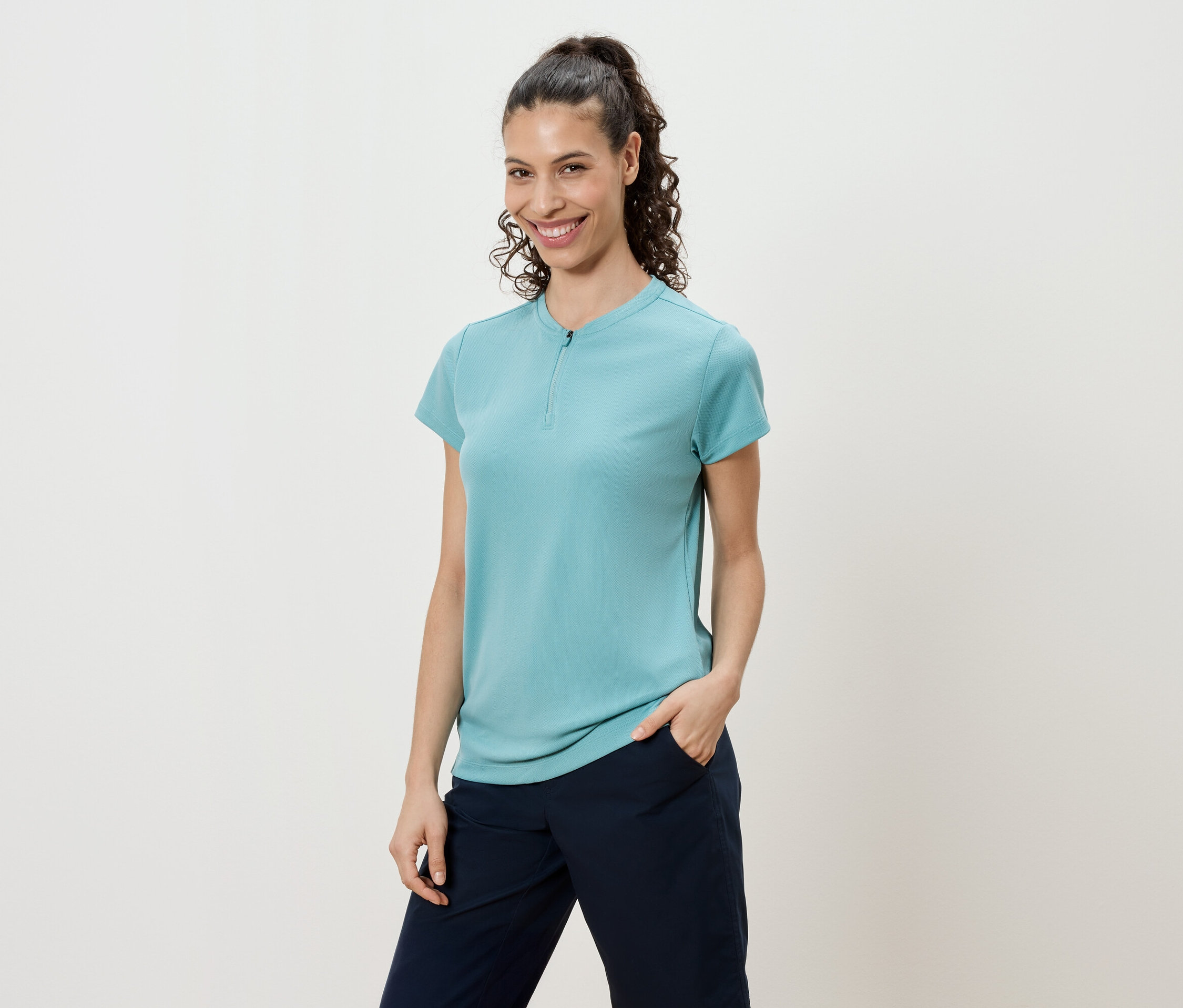 Žena s kudrnatými vlasy staženými do culíku má na sobě tyrkysové tričko a tmavě modré kalhoty.