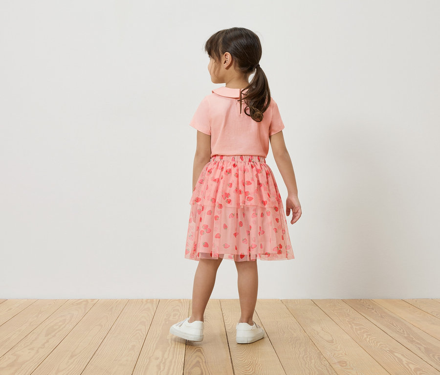 Dívka stojí zády ke kameře, má na sobě růžové tričko, růžovou tylovou sukni s potiskem jahod a bílé tenisky.